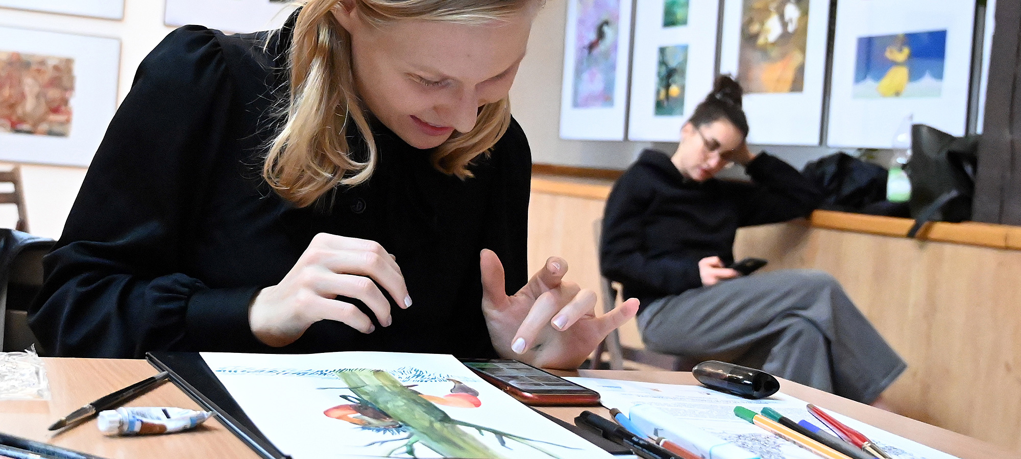 Uśmiechnięta młoda artystka- uczestniczka warsztatów podczas tworzenia ilustracji. Przed nią na stole rozłożone różne przybory malarskie.  