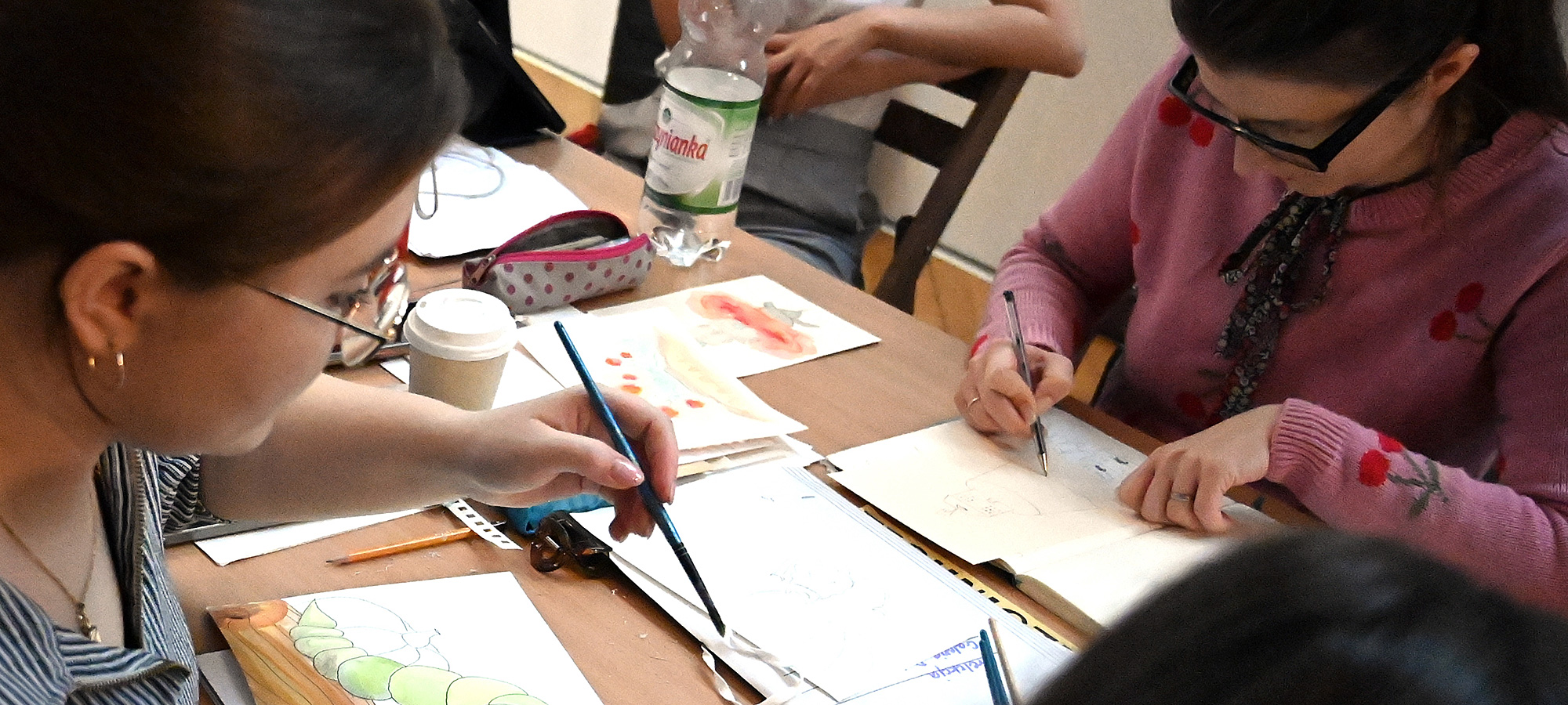 Dwie młode artystki- uczestniczki warsztatów podczas tworzenia ilustracji. Pomiędzy nimi na stole rozłożone różne przybory malarskie. Obie mają pędzle w dłoniach.